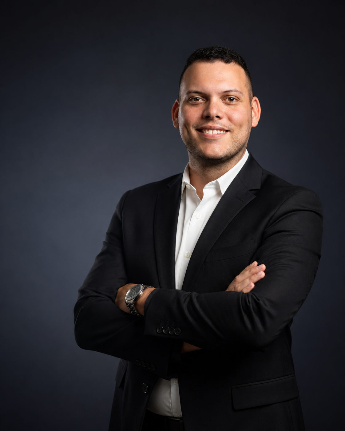 Retrato institucional de Samuca Trader em fundo escuro, vestindo terno preto e camisa branca, com braços cruzados e um sorriso confiante.